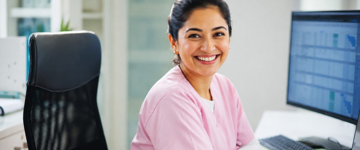 A smiling front-desk staff member at a clinic reception desk, with the schedule on screen.